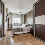 Modern bedroom featuring light wood flooring, dark wood sliding wardrobes on both sides, a stone accent wall behind a neatly made bed, and a bright window at the back.