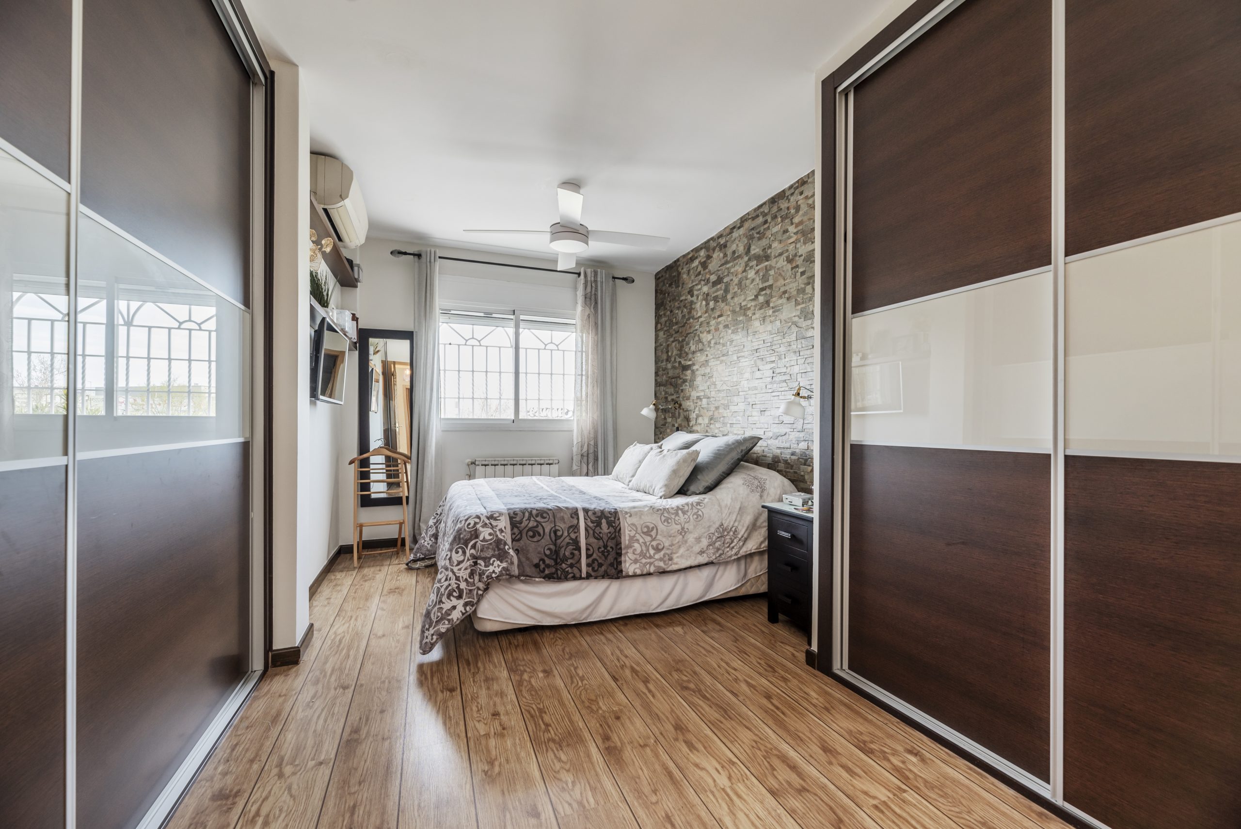 Modern bedroom featuring light wood flooring, dark wood sliding wardrobes on both sides, a stone accent wall behind a neatly made bed, and a bright window at the back.